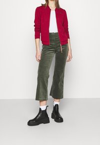 Veste zippée rouge avec un motif texturé, t-shirt blanc, pantalon large en velours côtelé vert olive, et bottines noires à semelle épaisse.