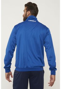 Giacca sportiva blu con colletto alto, maniche lunghe e polsini elasticizzati. Il retro presenta il logo "LEGEA" in bianco. Texture liscia.