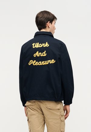 Άτομο που φοράει σκούρο μπουφάν με το κέντημα «Work And Pleasure» στην πλάτη και μπεζ παντελόνι cargo, με την πλάτη στραμμένη σε απλό φόντο.