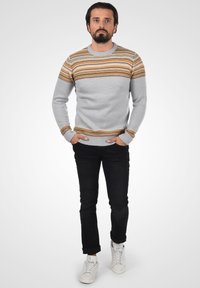 Grauer Pullover mit horizontalen orangen und gelben Streifen. Aus Strickstoff gefertigt, mit gerippten Bündchen und Saum. Getragen mit schwarzen Hosen und weißen Turnschuhen.