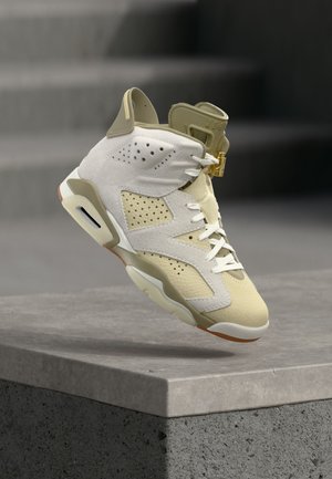 Air Jordan 6 tenisica u krem i bež nijansama, u zraku iznad betonske ivice, prikazujući perforiranu kožu i bijele vezice sa zlatnim kopčom.