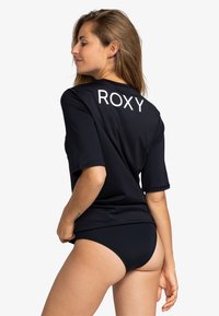 Navy blue Roxy swim shirt with short sleeves and white logo on the back, paired with matching black swim bottoms. Smooth texture.