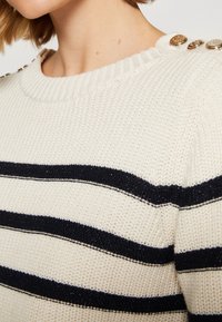Pull en maille crème avec des rayures horizontales noires et des boutons dorés décoratifs sur l'épaule, porté par une personne aux cheveux courts.