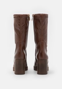 Bottes en cuir marron montantes avec une texture lisse, des bouts ronds et un talon en bois épais, dotées de fermetures éclair à l'arrière.