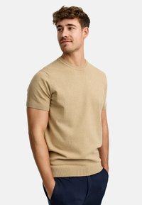 Suéter de punto beige de manga corta con escote redondo. Textura suave, puños y dobladillo acanalados, y corte holgado. Llevado con pantalones oscuros.
