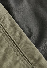 Des morceaux de tissu vert olive et gris foncé se superposent, mettant en valeur une texture lisse et des détails de couture visibles le long de la couture.