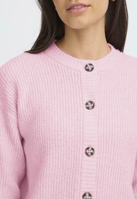 Cardigan roz tricotat cu guler rotund, având șase nasturi lucioși pe față și un țesut texturat pe margini. Material moale.