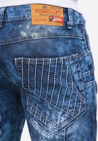 Jeans in denim blu sbiadito, con un'etichetta beige recante la dicitura "KOSMO·LUPO", cuciture dettagliate sulla tasca posteriore e una finitura testurizzata.