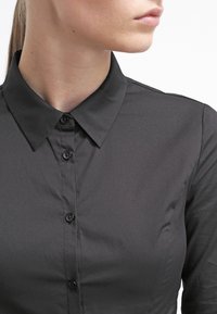 Camisa negra abotonada hecha de tela suave, con cuello clásico, botones pequeños y diseño ajustado, mostrando detalles mínimos de costura.