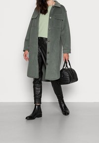 Grüne Fleecejacke mit Knopfverschluss und zwei Brusttaschen, kombiniert mit schwarzen Kunstlederhosen und Stiefeletten, hält eine schwarze Handtasche.