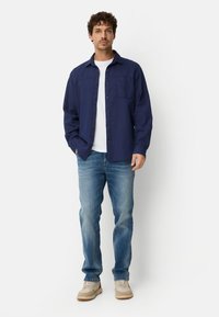 Man standing with hands in pockets wearing a navy button-up shirt over a white T-shirt, blue jeans, and beige sneakers against a white background.