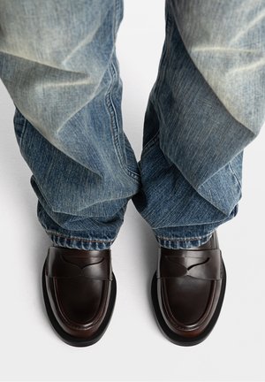 Person iført løstsiddende blå jeans og mørkebrune læderloafers, stående på en hvid overflade.