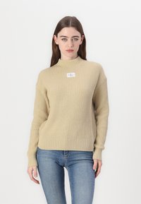 Beżowy sweter w prążki z wysokim kołnierzem i długimi rękawami, z małą naszywką z logo na klatce piersiowej. W połączeniu z jasnoniebieskimi dżinsami.