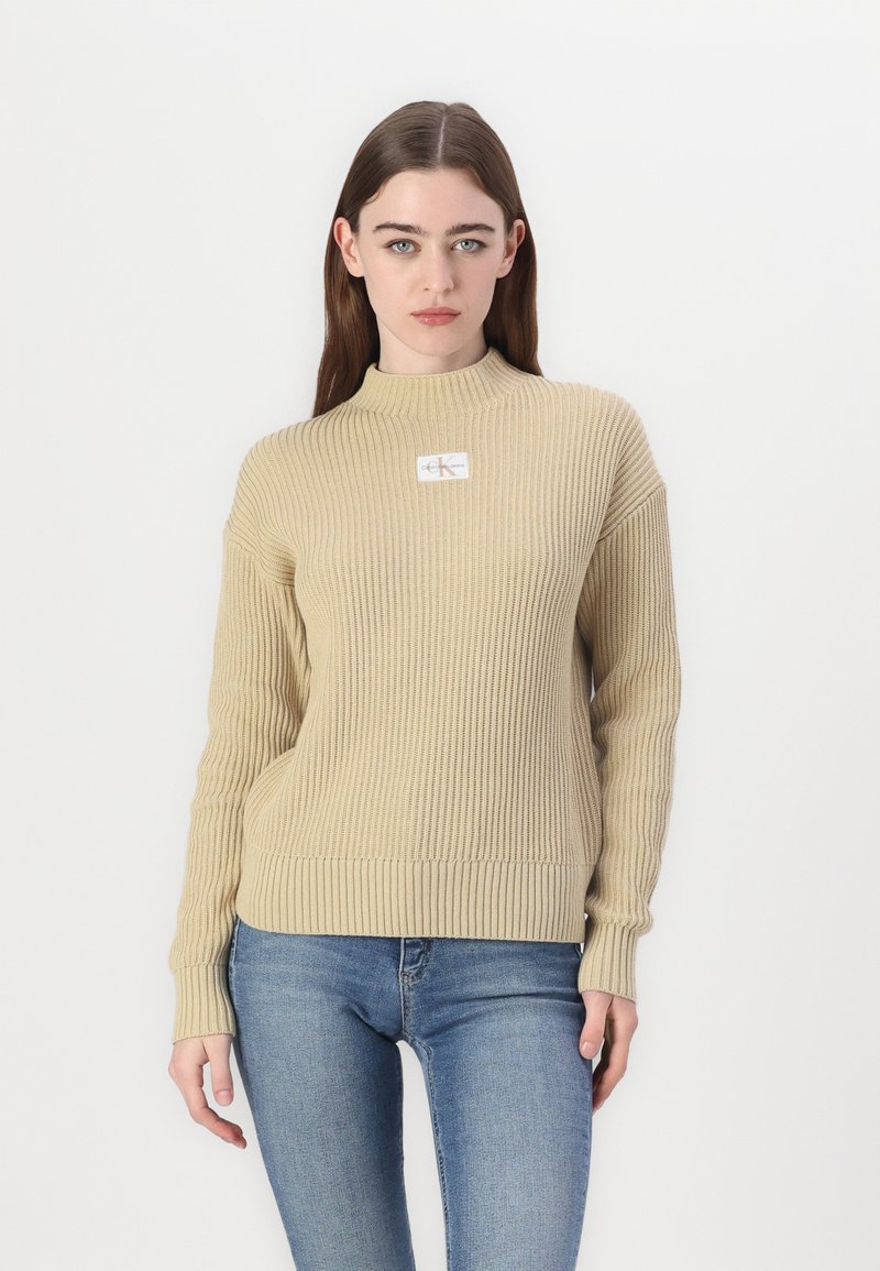 Beżowy sweter w prążki z wysokim kołnierzem i długimi rękawami, z małą naszywką z logo na klatce piersiowej. W połączeniu z jasnoniebieskimi dżinsami.