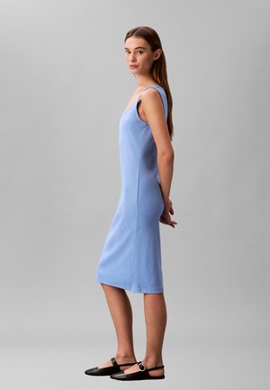 Tânără îmbrăcată într-o rochie midi bleu deschis fără mâneci și pantofi negri comozi, stând cu profilul și având mâinile la spate, în fața unui fundal gri simplu.