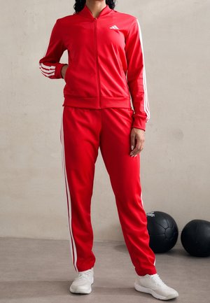 Persona che indossa una tuta rossa Adidas con strisce bianche e sneakers bianche, in piedi su un pavimento grigio vicino a due palle mediche nere.