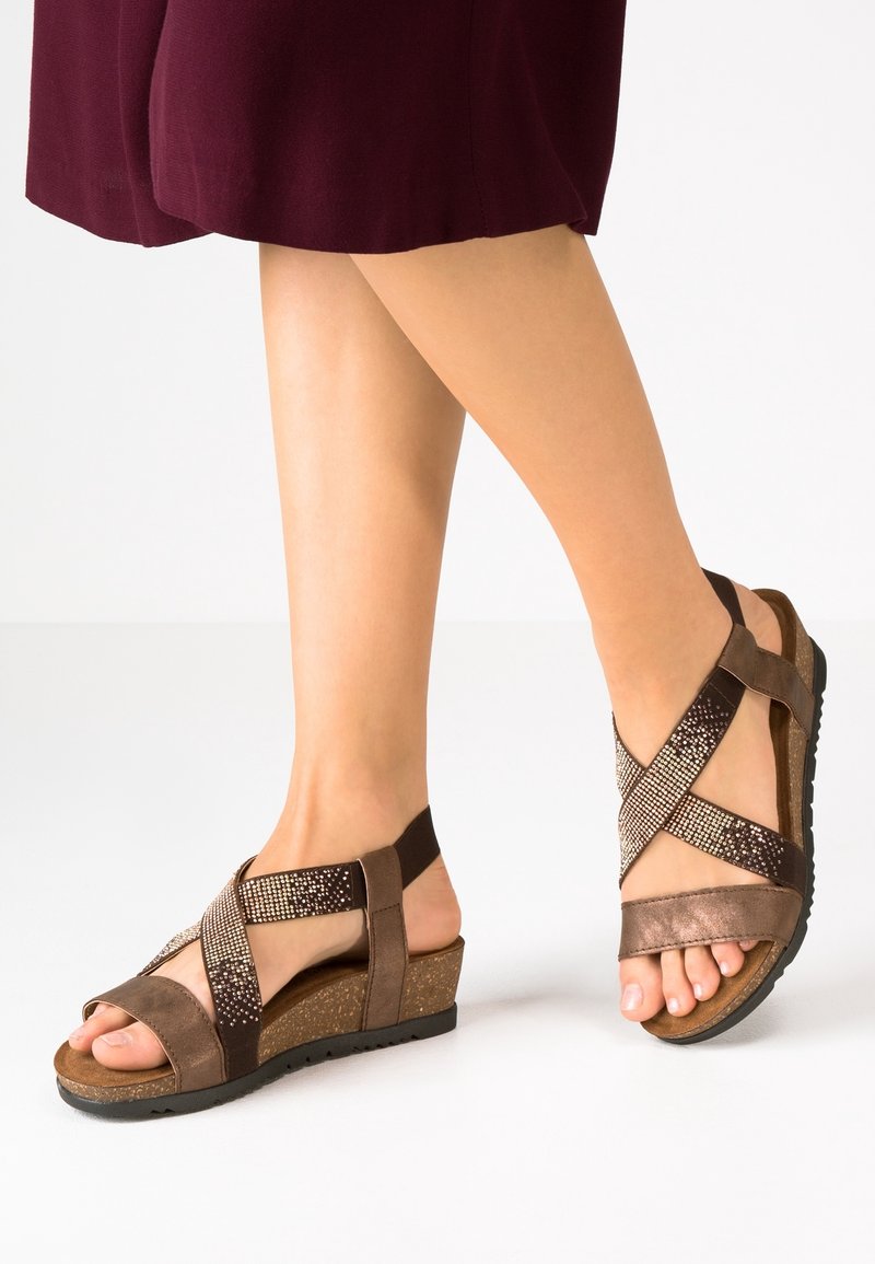 Sandales compensées brunes avec des lanières croisées, accents métalliques et une semelle texturée semblable au liège. Design à bout ouvert et bride arrière ajustable.