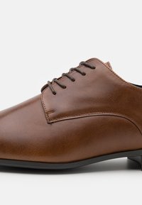Chaussure habillée en cuir marron avec une finition lisse, bout arrondi, accent cousu et lacets marron foncé, dotée d'un talon noir bas.