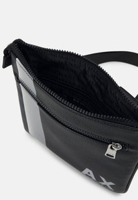 Borsa a tracolla nera con texture, striscia argentata, chiusura con zip e dettaglio del logo. Interno foderato con motivo a ripetizione del nome del marchio.