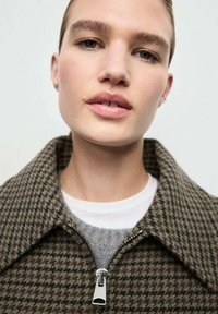 Chaqueta de houndstooth marrón con cremallera y gran cuello, superpuesta sobre un suéter gris y una camisa blanca. Tejido suave con detalles de cremallera brillante.