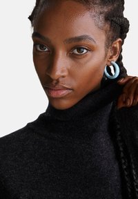 Schwarzer Rollkragenpullover aus strukturiertem Strick, kombiniert mit hellblauen Creolen. Das Model hat geflochtenes Haar und ein neutrales Make-up.