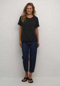 Camiseta negra de manga corta con cuello redondo, combinada con jeans recortados oscuros y chanclas negras, sobre un fondo neutro.