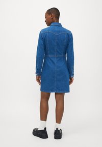 Denim obleka v srednji modri barvi z oprijetim pasom, dolgimi rokavi in gumbi na manšetah. Ima klasičen ovratnik in ukrivljen yoke na rami.