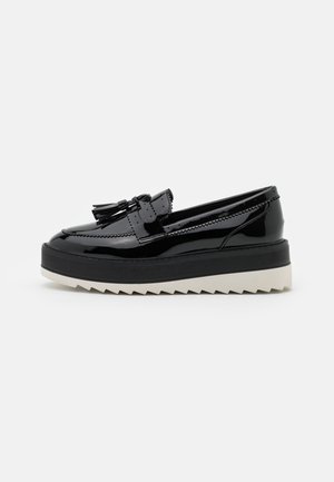Mocassin en cuir verni noir avec pompons, semelle intermédiaire noire épaisse et semelle extérieure blanche dentelée, présenté de profil sur fond blanc.
