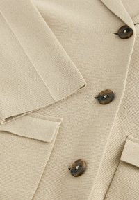 Béžový pletený cardigan s texturovaným povrchem, s tmavě hnědými knoflíky a přizpůsobeným střihem, včetně klop a kapsových klapek.