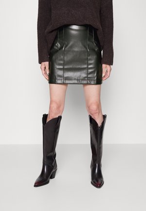 Stivali da cowboy in pelle nera con punte appuntite e altezze elevate, abbinati a una gonna in pelle verde scuro e a un maglione oversized marrone.