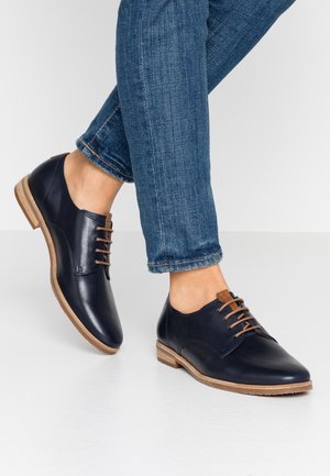 Scarpe in pelle blu navy con una finitura liscia, lacci marroni e dettagli color tan; abbinati a jeans blu, con una vestibilità slim e orli sfrangiati.
