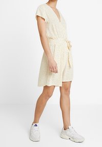 Robe courte jaune avec un décolleté en V et des manches courtes évasées, dotée d'une taille nouée et d'un motif à pois noirs. Assortie à des baskets blanches.