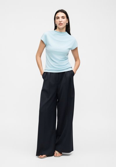 Γυναίκα στέκεται με τα χέρια στις τσέπες, φοράει ανοιχτό μπλε κοντομάνικη μπλούζα, φαρδιά μαύρη παντελόνα και μπεζ σανδάλια σε λευκό φόντο.