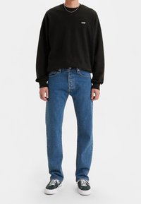 Sudadera negra con logo blanco, vaqueros azules de pierna recta y zapatillas negras con suelas blancas. Conjunto sencillo y casual con colores contrastantes.