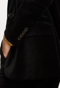Čierny sametový blazer s priliehavým strihom, gombíkovými detailmi na manžete a kontrastným zlatým lemom na rukáve.