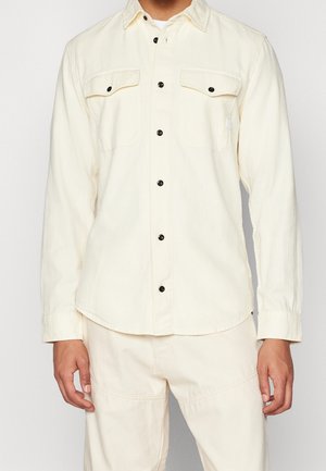 Mann trägt ein cremeweißes Hemd mit Knöpfen und zwei Brusttaschen sowie passende cremeweiße Hose vor einfarbigem Hintergrund.