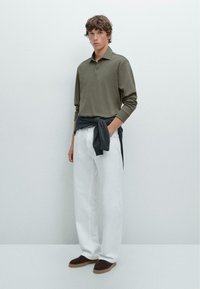 Khakifarbenes Langarm-Polo-Shirt, getragen mit weißen, locker sitzenden Hosen. Ein dunkler Pullover ist um die Taille gebunden und wird mit braunen Wildlederschuhen kombiniert.
