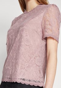 Femme portant un haut en dentelle florale rose poussiéreux à manches courtes avec des bords festonnés sur une couche unie, associé à un bas noir.