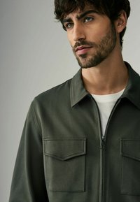 Chaqueta verde oliva con cremallera, dos bolsillos en el pecho, cuello, textura estructurada y un interior suave. Camiseta blanca de cuello redondo debajo.