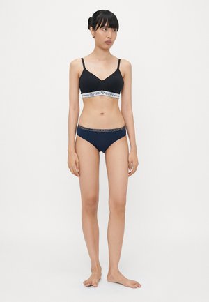 Melnā bezvadu bikini augšdaļa ar baltu zīmolu apakšmalu, apvienota ar atbilstošām melnām bikini apakšdaļām, kas iezīmē elastīgas akcentus un gludu tekstūru.
