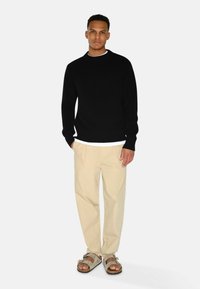Pulover negru cu model canalat, pantaloni bej plisati și sandale gri. Design simplu, cu gât rotund și croială dreaptă. Fără modele vizibile.