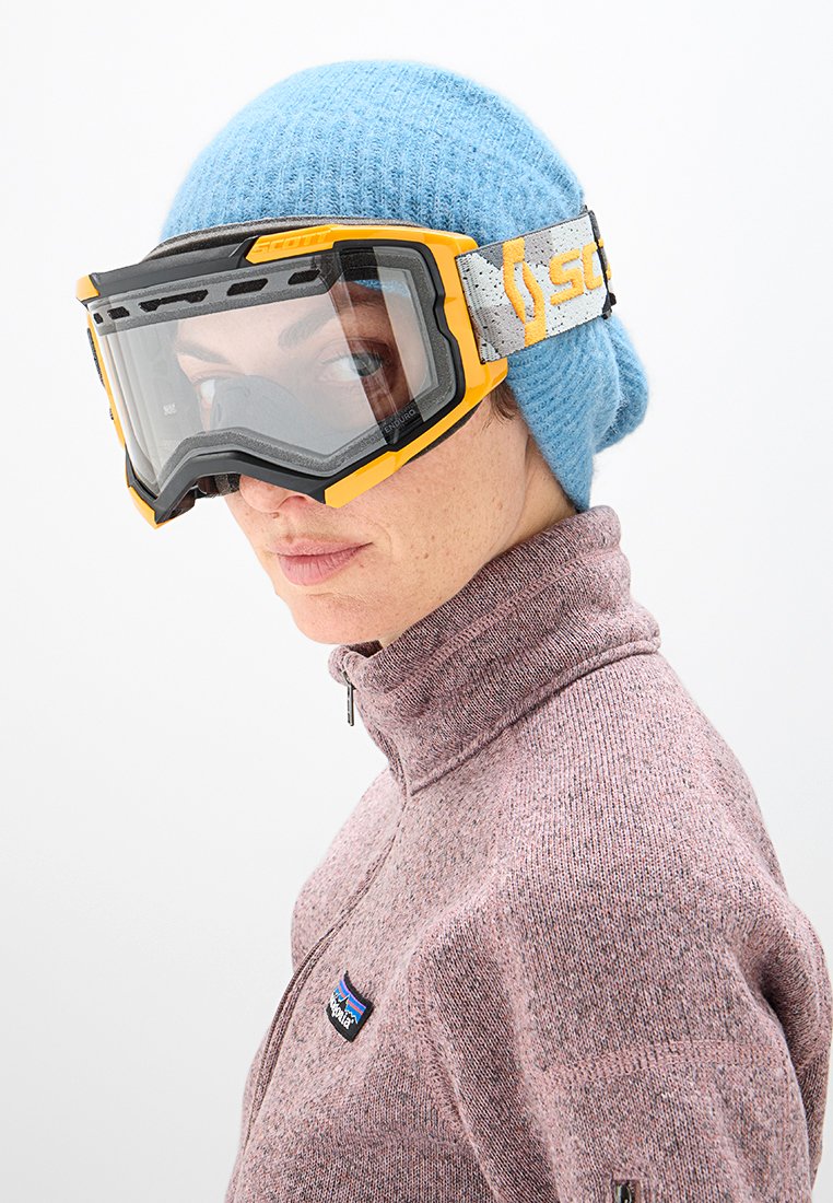 Gafas de esquí con lentes transparentes y acentos amarillos, llevadas sobre un gorro de punto azul, combinadas con un suéter de cremallera en tono rosa jaspeado.