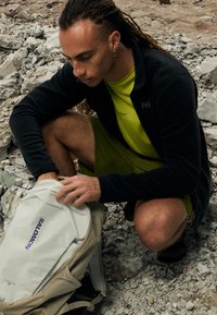 Bež nahrbtnik z logotipom "SALOMON", narejen iz trpežnega blaga. Površina je gladka. Posameznik kleči na skalnati podlagi, oblečen v črno flis jakno in svetlo zeleno majico.