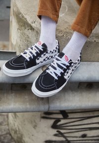 Black and white canvas sneakers with a checkerboard design, white laces, red logo patch, and contrasting stitching.