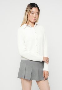 Vit, mjuk stickad cardigan med krage, prydd med pärlknappar och långa ärmar, i kombination med en grå plisserad kjol.