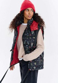 Czarna pikowana kamizelka w kwiaty z czerwonymi akcentami, lekka tkanina, zamykane na zamek kieszenie i paski na ramiona, noszona na beżowym dzianinowym swetrze.