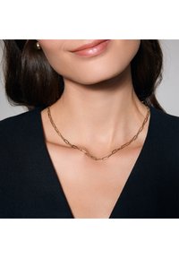 Złoty naszyjnik łańcuszkowy z owalnym wzorem ogniw. Noszony blisko szyi, wyeksponowany na ciemnym tle.