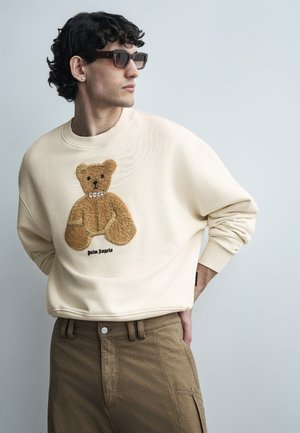 Νεαρός με σγουρά μαλλιά που φοράει σκούρα γυαλιά ηλίου, κρεμ φούτερ με σχέδιο αρκουδάκι και καφέ παντελόνι, κοιτάζει προς τα πλάγια.