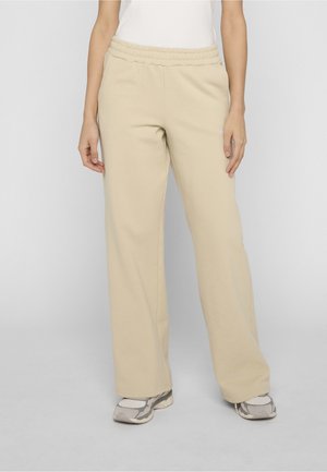 Kvinne kledd i beige vide bukser og hvite joggesko, stående mot en ensfarget, lys grå bakgrunn.