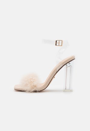 Beżowe sandały na wysokim obcasie z przejrzystym paskiem w kostce, z puszystym, jasnym pomponem na palcu. Przejrzysty blokowy obcas.